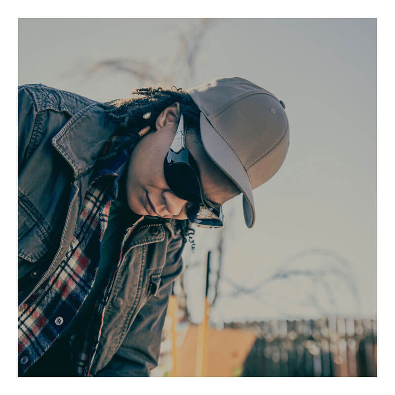 A person in a khaki cap, plaid shirt, jacket, and MCR Safety Law OTG Safety Glasses UV-AF Anti-Fog OG1 looks down outdoors on a sunny day, with a blurred fence and branches in the background.