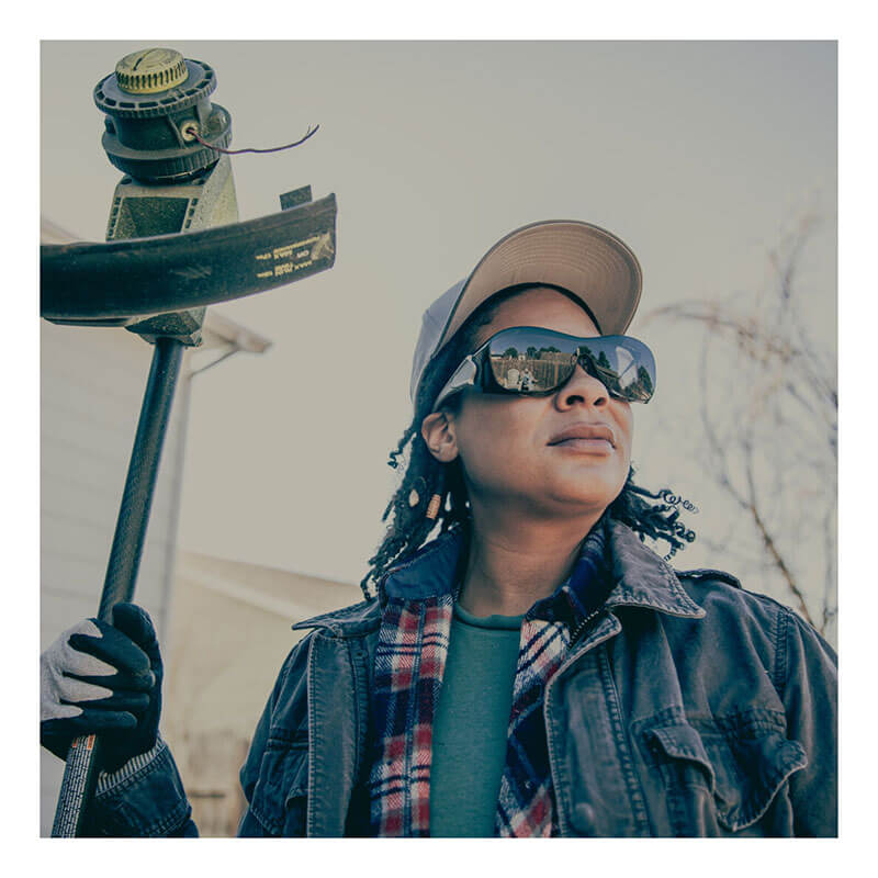 A person wearing MCR Safety Law OTG Safety Glasses UV-AF Anti-Fog OG1, a hat, gloves, and a plaid shirt under a jacket holds a string trimmer outdoors on a clear day, looking into the distance.