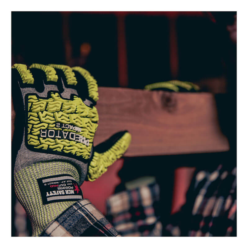 A person wears MCR Safety Predator Hi-Vis Mechanics Gloves w/HyperMax PD6952 (12 pairs) in green and black, holding a wooden plank as the cut-resistant gloves stand out in the dimly lit workshop.