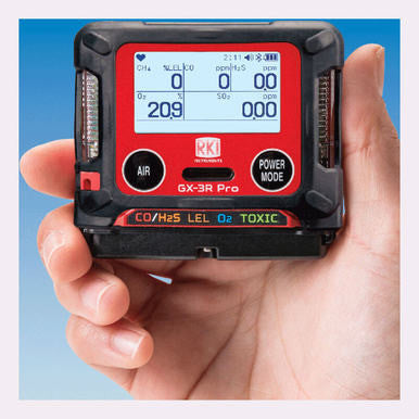 A hand holding the RKI GX-3R Pro Gas Confined Space Monitor, a stylish personal device from RKI featuring wireless communication and a digital display indicating CO, H2S, LEL, and O2 levels. The monitor showcases a black and red design with buttons labeled "Air" and "Power Mode.