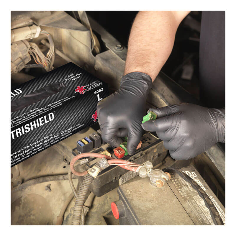 A person wearing MCR Safety NitriShield Stealth Extra Gloves 6062 is working on a car engine, holding a green fuse near a fuse box. A box of MCR Safety NitriShield gloves is in the foreground.