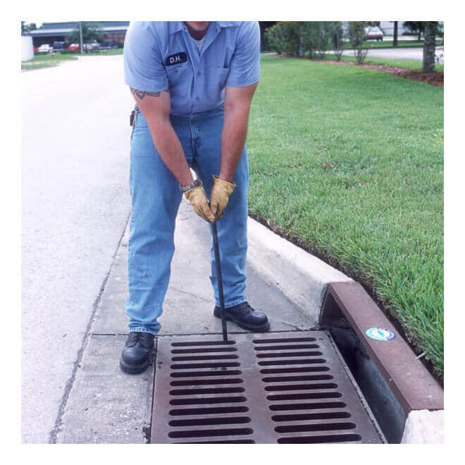 A person in a blue uniform and safety gear uses the UltraTech Grate Hook 9235 to lift a storm grate on a street next to a grassy area.