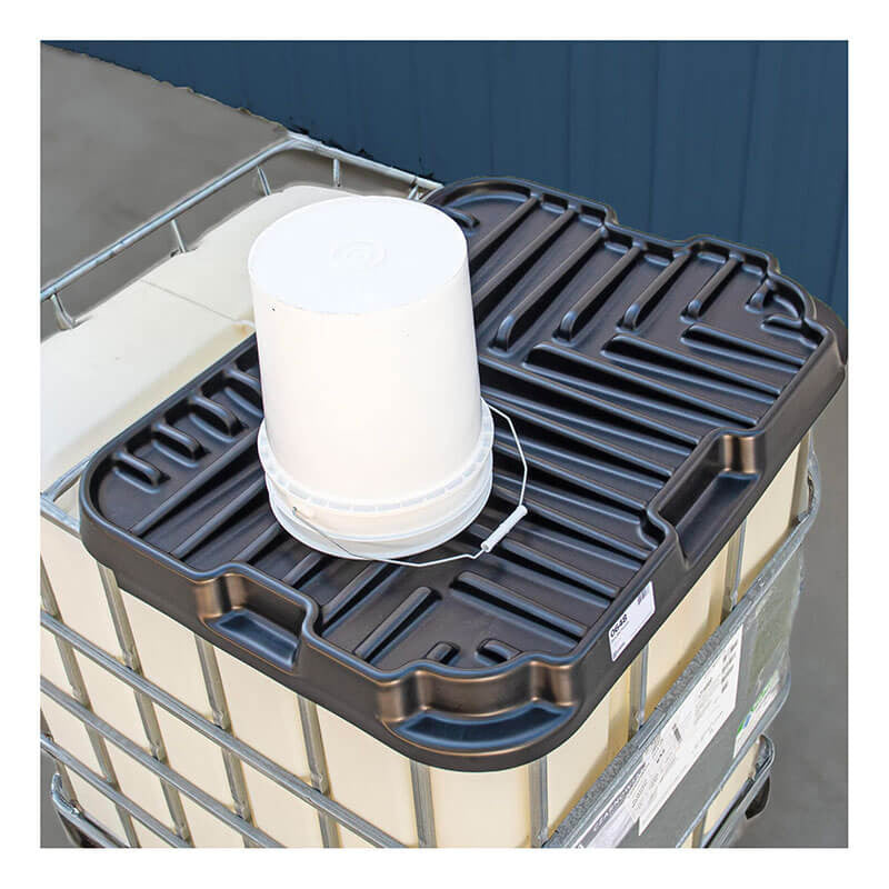A white plastic bucket rests on a black, grooved plastic lid that covers a large metal container, resembling the UltraTech IBC Funnel 0648 by UltraTech International, Inc. The setting appears industrial, featuring a blue wall in the background.