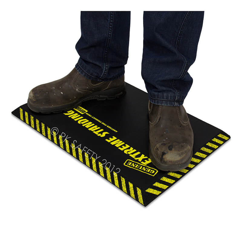 A person wearing brown work boots stands on a black Working Concepts ErgoKneel Extreme Standing Mat 14 x 21 in., featuring yellow caution stripes and the text "EXTREME STANDING" in bold, while utilizing an anti-fatigue mat for enhanced comfort.