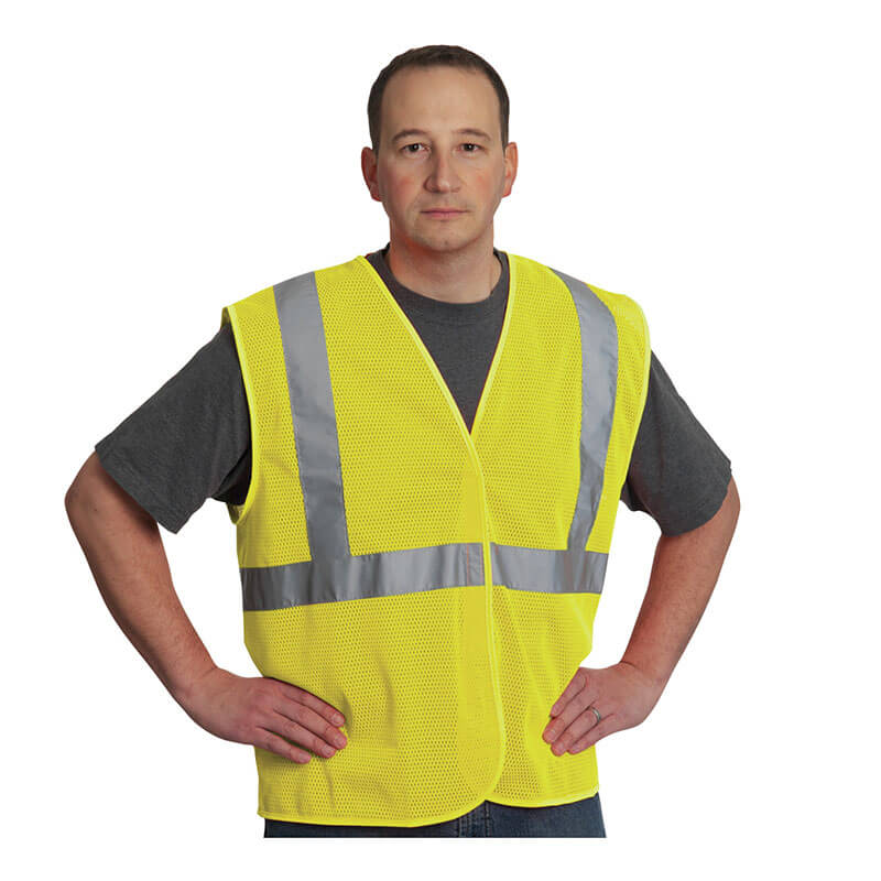 A man in a gray t-shirt and a PIP Type R Class 2 Two-Tone Value Mesh Vest 302-MVG stands with his hands on his hips against a white background. This PIP vest offers ANSI-compliant visibility and comfort with reflective silver stripes.