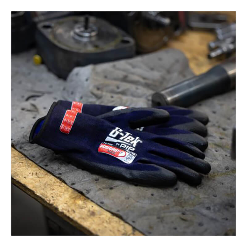 A pair of black PIP G-Tek PosiGrip MAX Nylon/Spandex Glove 34-188 rests on a grey mat on a cluttered workbench, surrounded by metal tools and machinery parts.