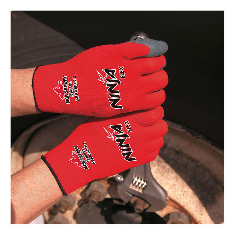 Wearing MCR Safety Memphis Ninja Flex Glove N9680, a person uses a wrench to tighten a bolt on a large vehicle wheel. The red gloves feature text highlighting excellent abrasion resistance.