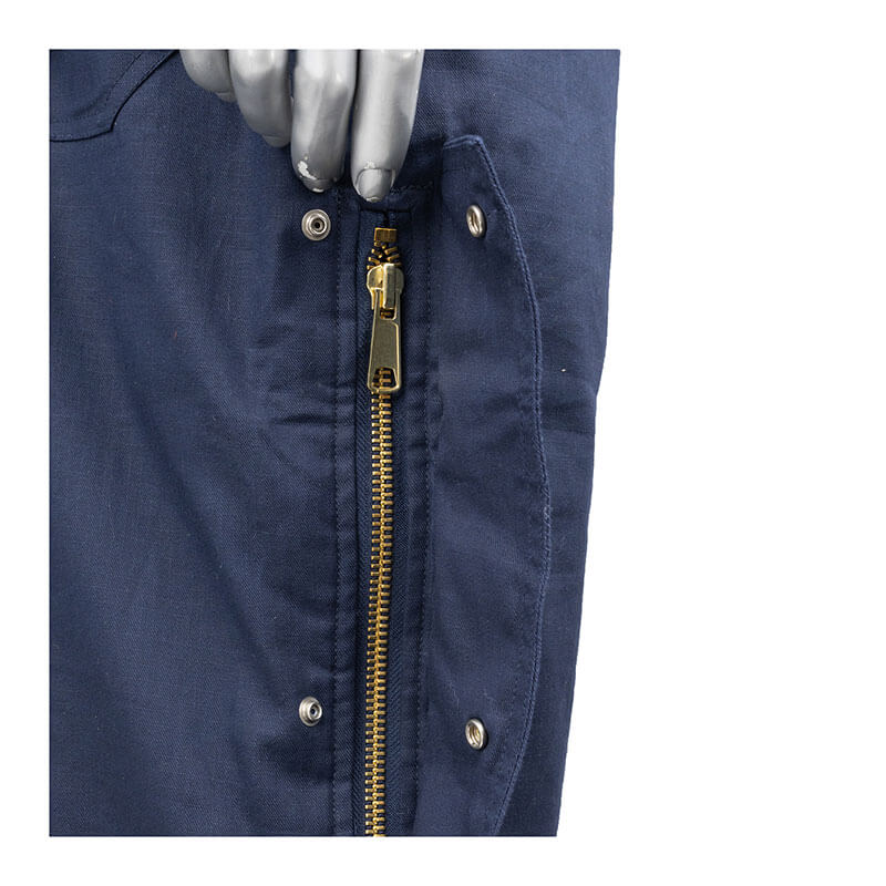 A close-up of the PIP AR/FR Dual Certified Insulated Bib Overall 385-FRBO in blue shows a gold zipper and metallic snap buttons, noted for flame resistance. A mannequin hand next to the zipper highlights these fastening features typical of insulated bib overalls.