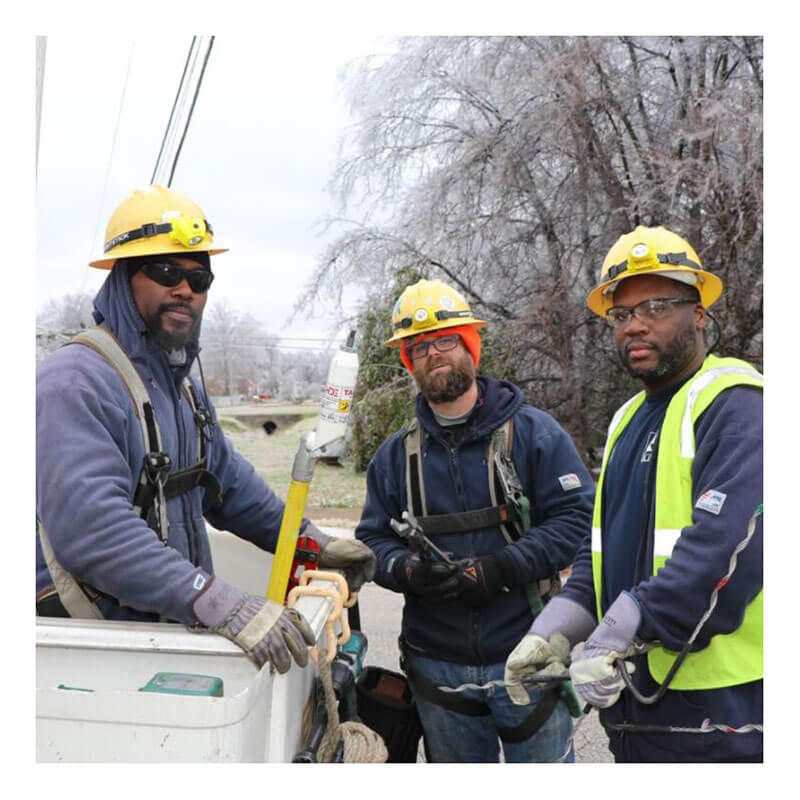 Three utility workers wearing safety helmets, harnesses, and MCR Safety Mustang Grain Leather Palm Gloves 1935 (12 pairs) stand outdoors near power lines and iced trees, posing for a photo while holding tools and equipment.