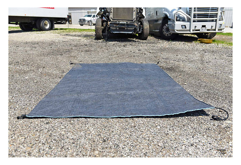 An UltraTech Spill Diaper 0275, produced by UltraTech International, Inc, is laid out on a gravel surface near two parked trucks. The foreground emphasizes this absorbent spill containment tarp, while the trucks are blurred in the background under a clear sky, underscoring its importance in environmental protection.