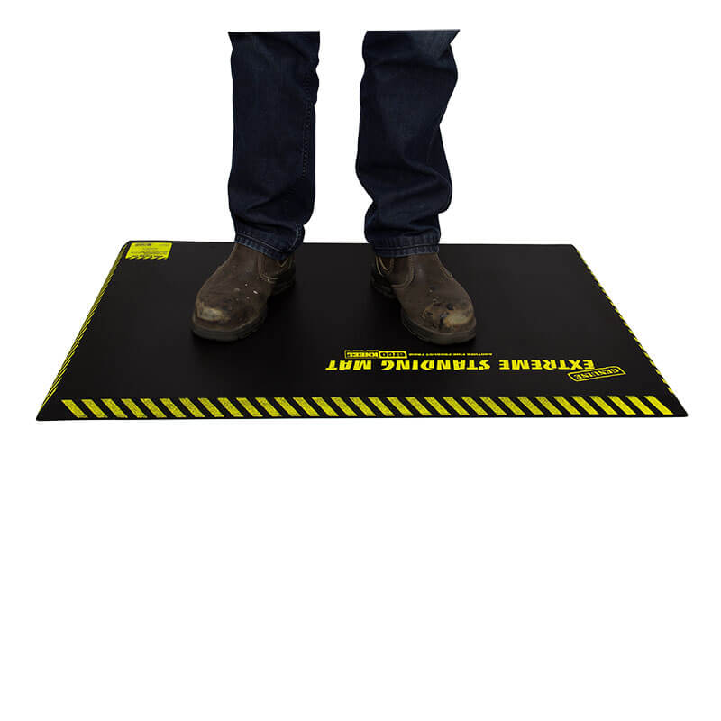 A person in brown work boots and blue jeans stands on a Working Concepts ErgoKneel Extreme Standing Mat 5032, featuring a black surface with yellow caution stripes. This anti-fatigue mat, made from closed cell foam pad material, provides comfort for long periods of standing while working.