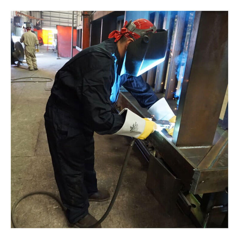 Wearing an MCR Safety FR Contractor Coverall Navy CCMN and a welding helmet, a worker welds metal in an industrial workshop with sparks flying—a standard safety practice in the oil and gas industry. Another employee is visible in the background.