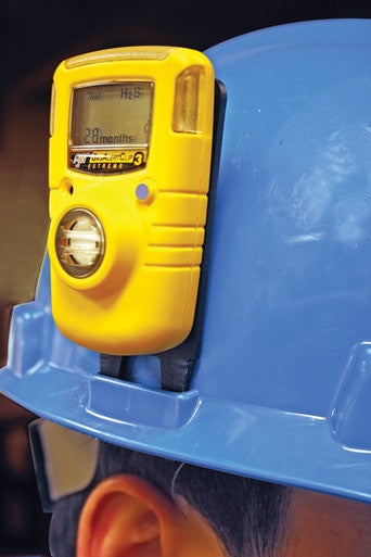 A hard hat, in blue, is worn by a worker with a yellow gas detector securely attached using the BW Hard Hat Clip GA-HC-1 from PK Safety Supply. The device's screen shows "28 months" and an "H2S" level for efficient gas detection in the field.