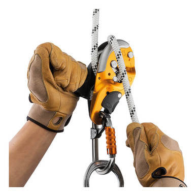 A person wearing tan gloves is adjusting a rope through a Petzl I'D S Descender Self-Braking D020AA00, which features an orange and black design. This device, used in technical rescue operations, is attached to a climbing carabiner. The rope is white with black speckles.