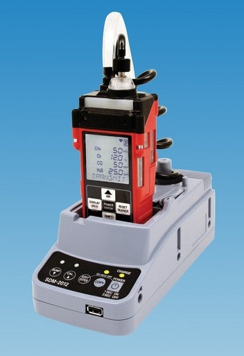 A red PK Safety Supply portable gas monitor sits on a gray RKI SDM-2012 Calibration Station for GX-2012, set against a blue background. The device's screen displays digital readings, with various buttons and connectors clearly visible on the base unit.