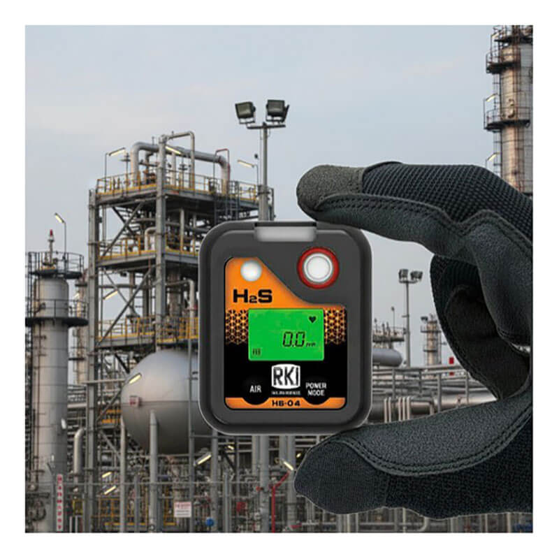A gloved hand holds an RKI Instruments HS-04 Series H2S Single Gas Monitor 73-0063 with a digital display reading “0.0,” set against an industrial facility featuring metal structures and tanks in the background.