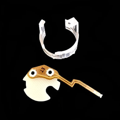 A small, intricate electronic component featuring a gold circuit pathway and white elements is displayed against a black background. Above it, there is a clear plastic clip-like holder, resembling those used for the PK Safety Supply BW Micro Clip XT/XL Replacement Flex PCB MC2-FPCB1.