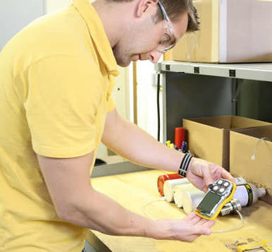 A person wearing safety glasses and a yellow shirt examines a BW Honeywell Gas Monitor at a workbench filled with tools and boxes, ensuring the device’s precision with expert calibration services from PK Safety Supply.