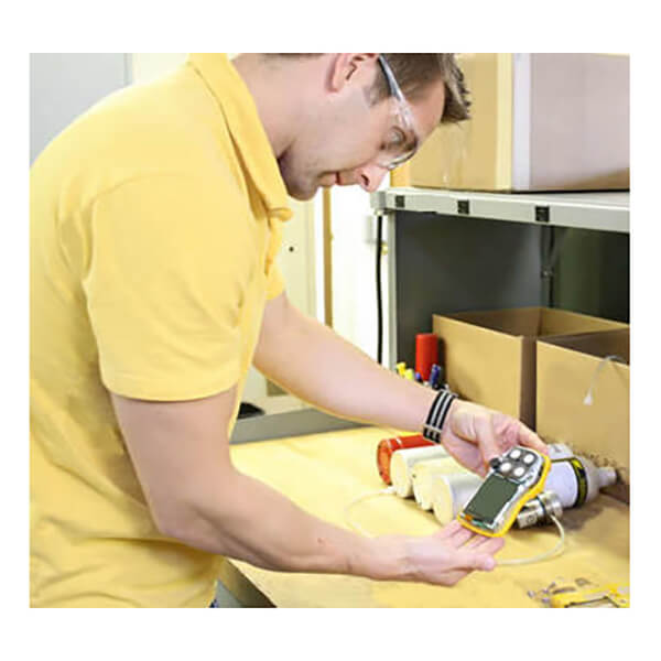 A person wearing safety glasses and a yellow shirt examines a BW Honeywell Gas Monitor at a workbench filled with tools and boxes, ensuring the device’s precision with expert calibration services from PK Safety Supply.
