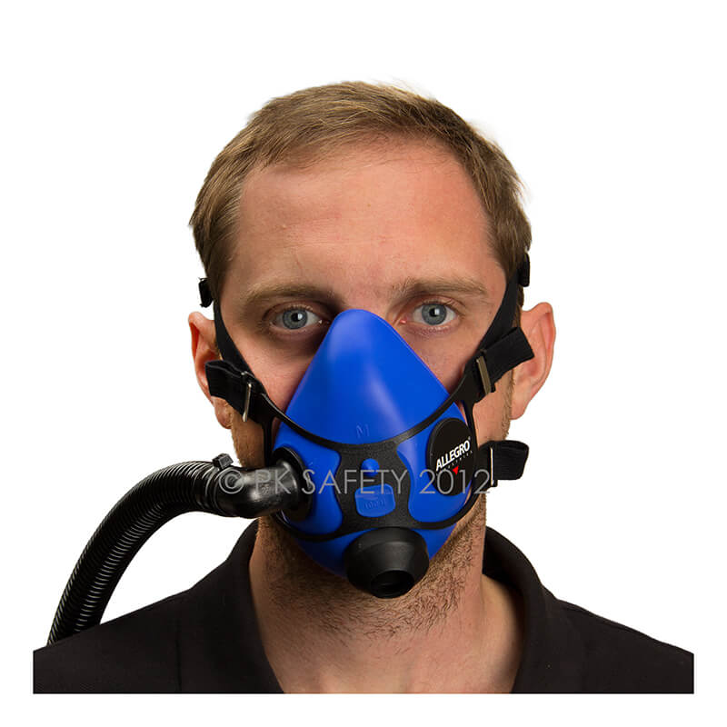 The man is confidently facing forward, dressed in a black shirt, while wearing the Allegro Half Mask Constant Flow Airline Respirator 9920 by PK Safety Supply. This blue mask features black straps and a side hose attachment for continuous airflow. The silicone facepiece covers his nose and mouth to ensure full protection.
