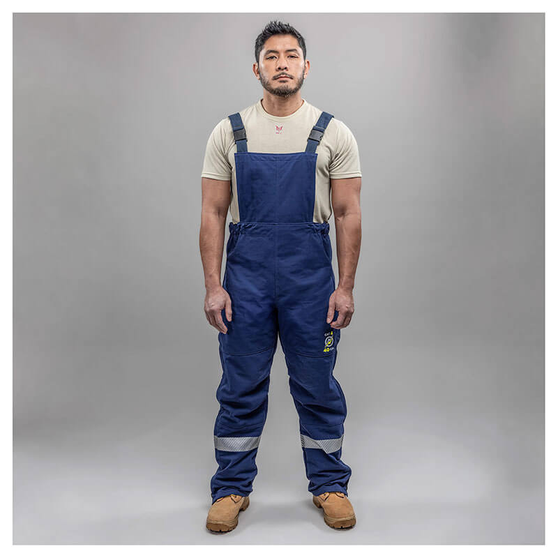 A man stands against a gray background wearing a light T-shirt, tan work boots, and NSA Enespro ArcGuard Performance 40 Cal Arc Flash Bib ARC40B coveralls with reflective leg bands for enhanced arc flash protection.