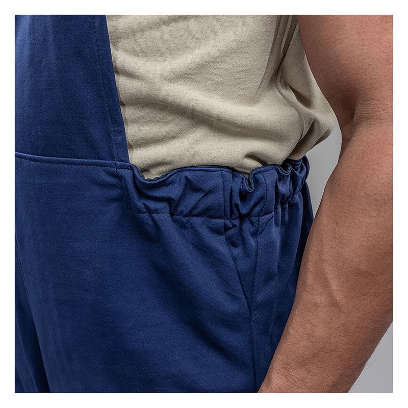 A close-up of a person wearing a beige shirt and blue NSA Enespro ArcGuard Performance 40 Cal Arc Flash Bib ARC40B, with their right hand in the pocket for arc flash protection.