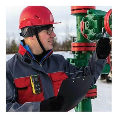 A worker with a red hard hat and glasses inspects a green and red valve outdoors, holding a clipboard. Clipped to his jacket is the Honeywell BW Flex4 4-Gas Detector by BW Technologies, monitoring gas hazards as snow blankets the ground.