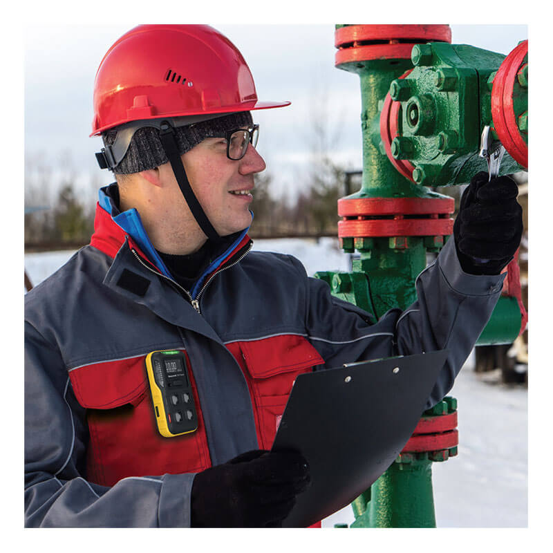 A worker with a red hard hat and glasses inspects a green and red valve outdoors, holding a clipboard. Clipped to his jacket is the Honeywell BW Flex4 4-Gas Detector by BW Technologies, monitoring gas hazards as snow blankets the ground.