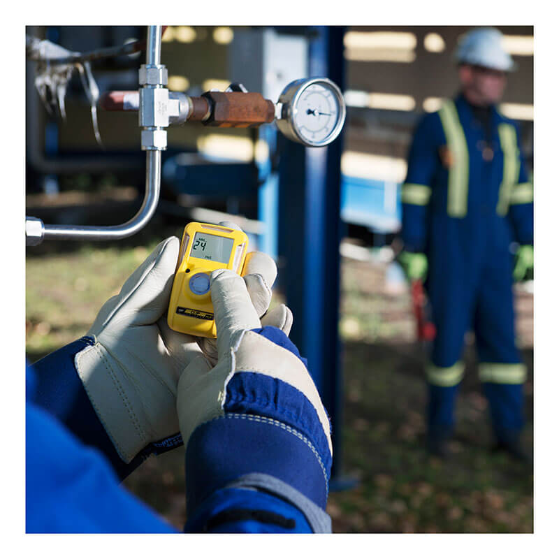 A worker in a blue protective suit and safety gloves uses the BW Technologies by Honeywell BW Clip 2 Year H2S Single Gas Detector (BWC2-H) near industrial equipment, with another worker in safety gear blurred in the background.
