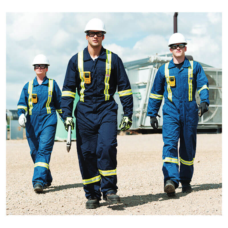 Three industrial workers in blue coveralls and vests, wearing BW Technologies by Honeywell BW Clip4 4-Gas Detector BWC4-Y-N, walk outdoors on gravel with a large wrench. Industrial equipment and a cloudy sky are seen in the background.