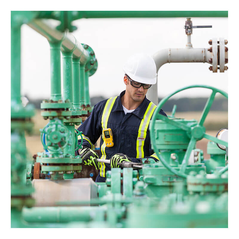 A worker in a white hard hat, safety glasses, gloves, and a reflective vest adjusts green pipes outdoors. A BW Technologies by Honeywell BW Clip4 4-Gas Detector BWC4-Y-N is clipped to the worker's vest.