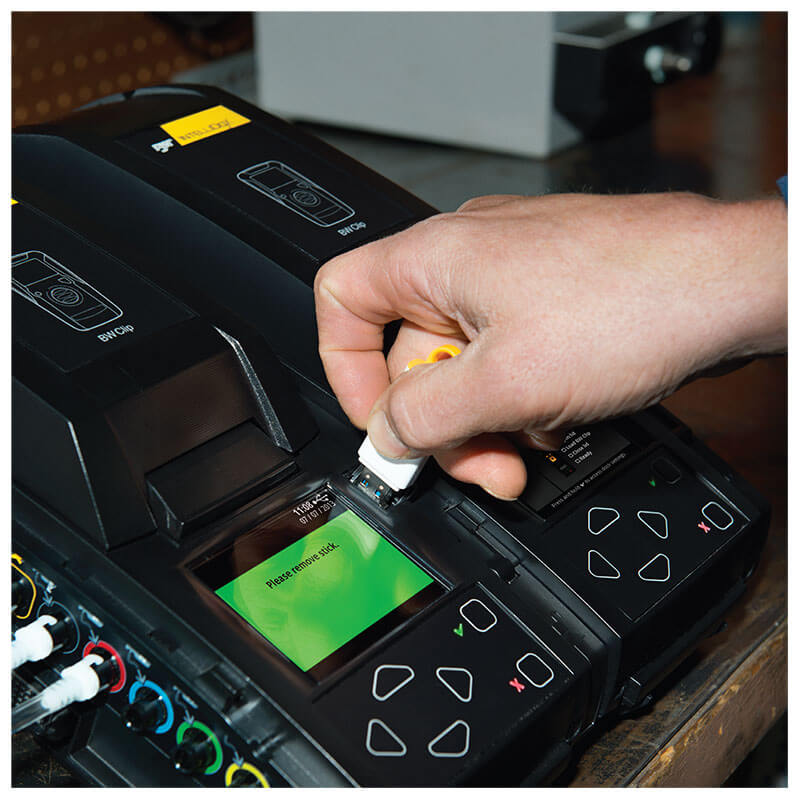 A hand inserts a USB flash drive into a black BW IntelliDoX Docking Station DX-CLIP by BW Technologies (Honeywell), featuring a green display and buttons as part of an advanced instrument management system, with another unit nearby.