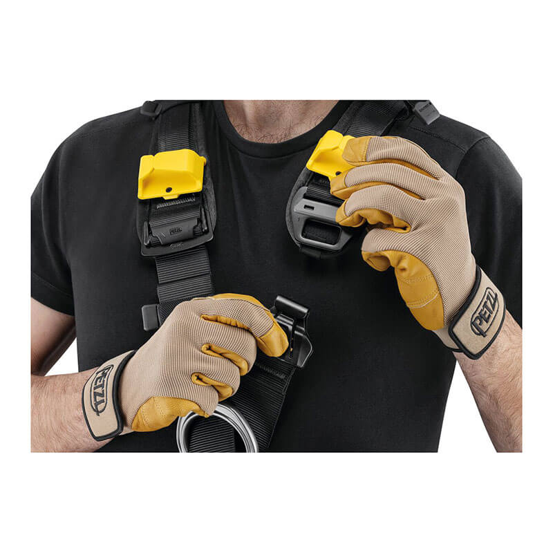 A person wearing yellow and beige gloves and a black shirt is adjusting a Petzl ASTRO International Harness (Model: C083BB0_) with its distinctive yellow features, exemplifying the precision of rope access technicians.
