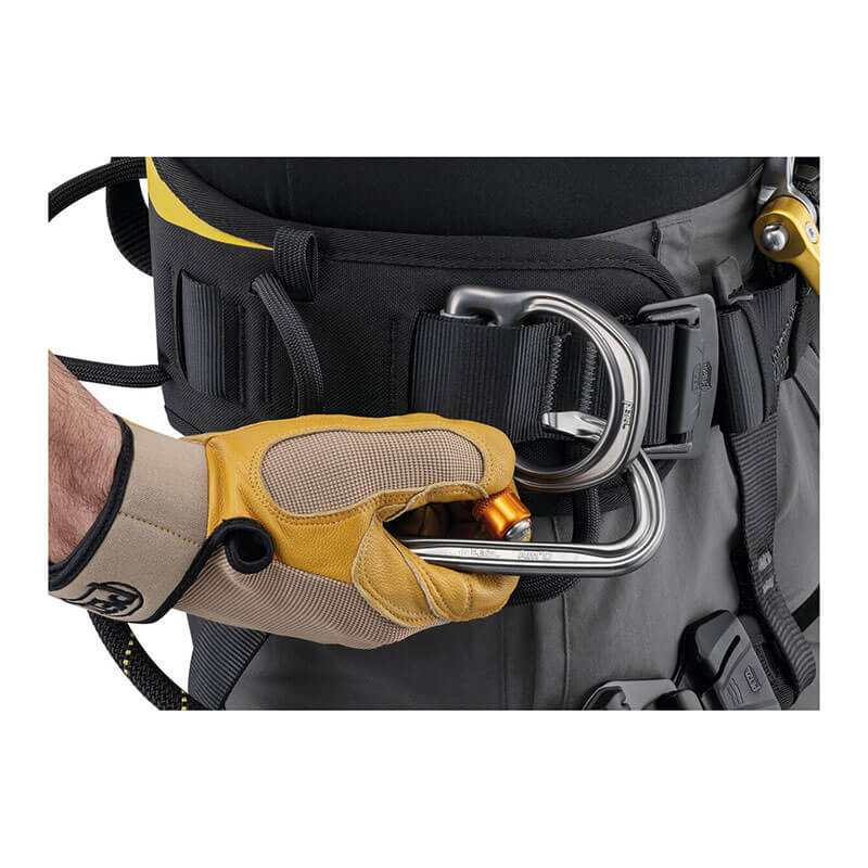 A gloved hand secures a metal carabiner to the Petzl ASTRO International Harness C083BB0_, a black and gray model favored by rope access technicians, adjusting or locking it with precision.