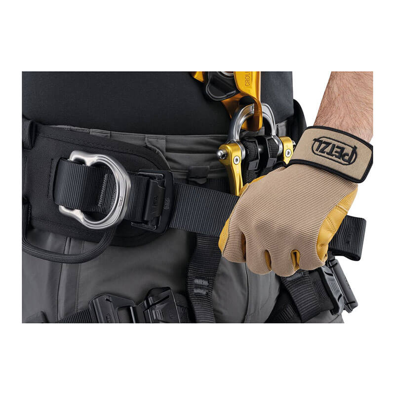 A rope access technician sporting black and gray work pants is seen adjusting a Petzl ASTRO International Harness C083BB0_ using yellow and black gloves. They grip a buckle, with an additional metal ring and clip visible on the belt, emphasizing the equipment and hands.