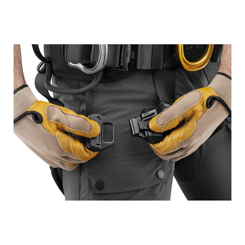 A person, wearing yellow and beige gloves, expertly connects two black buckles on a Petzl ASTRO International Harness C083BB0_, designed for rope access technicians. Dressed in dark clothing, the individual focuses on the task against a plain white background.