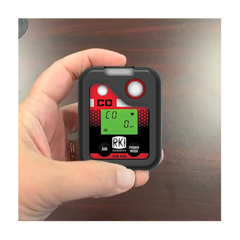 A hand holds an RKI Instruments CO-04 Series CO Single Gas Monitor 73-0065 with a digital display reading "CO 0 ppm" and "RKI," set against a dark wooden surface.