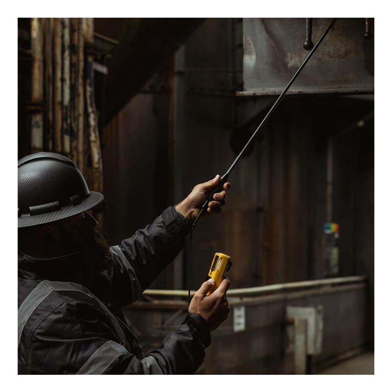 Wearing a hard hat and safety jacket, an individual inspects an industrial duct in a dimly lit setting using the WatchGas Fixed Carbon Fiber Probe with Teflon Insert from WatchGas to meticulously examine the area for gas detection.