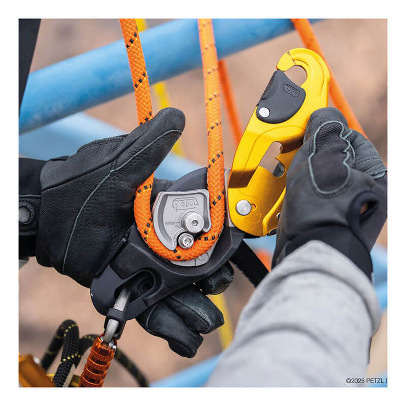 A close-up of gloved hands using the Petzl RIG Compact Descender D021_ _00 to attach an orange rope to its AUTO-LOCK system with yellow and black parts, set against a blue metal structure in the background.