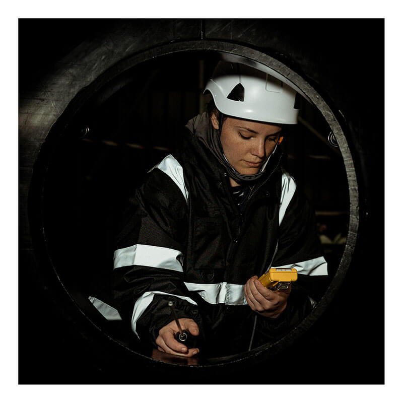 A worker in a hard hat and reflective jacket uses the WatchGas SST4-PUMP 4-Gas Detector from WatchGas inside a large cylindrical pipe, concentrating on their task in a dimly lit industrial setting.