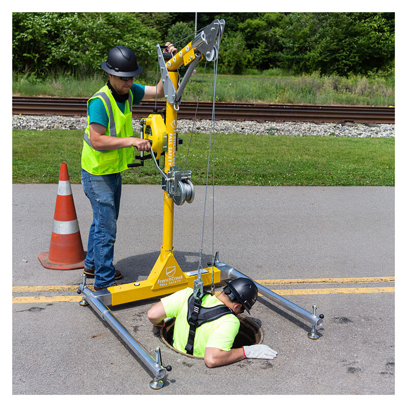 Two workers in safety gear use the French Creek Production Portable Davit System Winch Mounting Bracket PD4 near a manhole; one operates the winch while the other enters a confined space. A traffic cone and railroad tracks can be seen in the background.