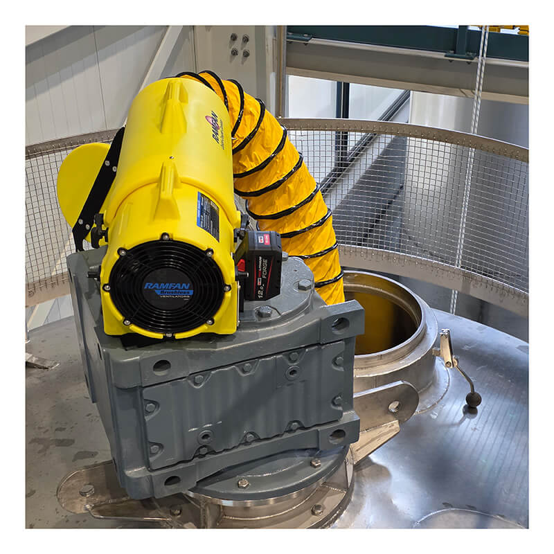 A Euramco RAMFAN UB20 18VDC industrial ventilator with a Quick-Couple Canister and flexible duct is connected to a gray metal opening on top of a large tank, delivering confined-space ventilation within the facility and enclosed by a metal safety railing.