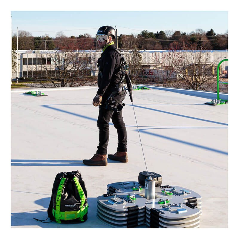 A worker in safety gear uses the Frontline Commando Counterweight Fall Protection System CO2NN by Frontline Fall Protection. Nearby is a green and black backpack, with trees and buildings in the background.