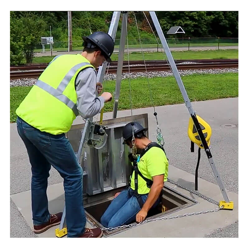 Two workers in safety gear prepare for confined space entry; one operates the French Creek Production 9' Tripod 3-Way Rescue Unit and Winch S50G-M9, while the other, harnessed, sits at an open hatch with railroad tracks and greenery behind them.