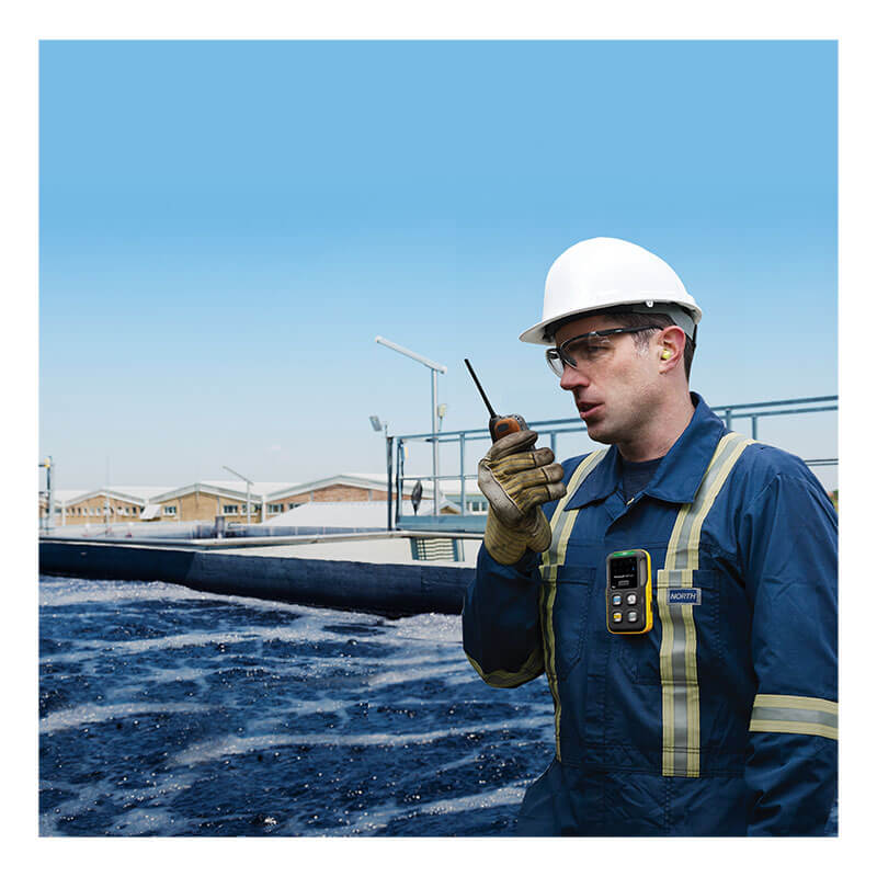A worker in a hard hat and protective gear stands by a large outdoor industrial water tank, using a walkie-talkie and wearing the BW Technologies by Honeywell BW Flex4 Gas Detector (%LEL(F), O2, H2S, CO), with blue sky and buildings behind.