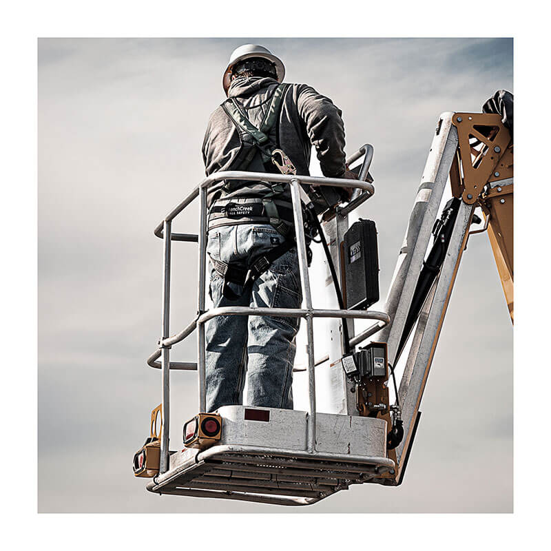 A construction worker in a French Creek Production STRATOS Construction Harness 22850B and helmet stands on an elevated cherry picker platform, working at height beneath a cloudy sky.