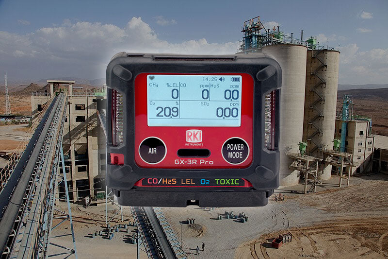A close-up of the RKI Instruments GX-3R Pro 5 Gas Confined Space Monitor with Wireless Communication appears over an industrial plant with silos and conveyors in a dry, mountainous area. The monitor's screen displays readings for multiple gases.