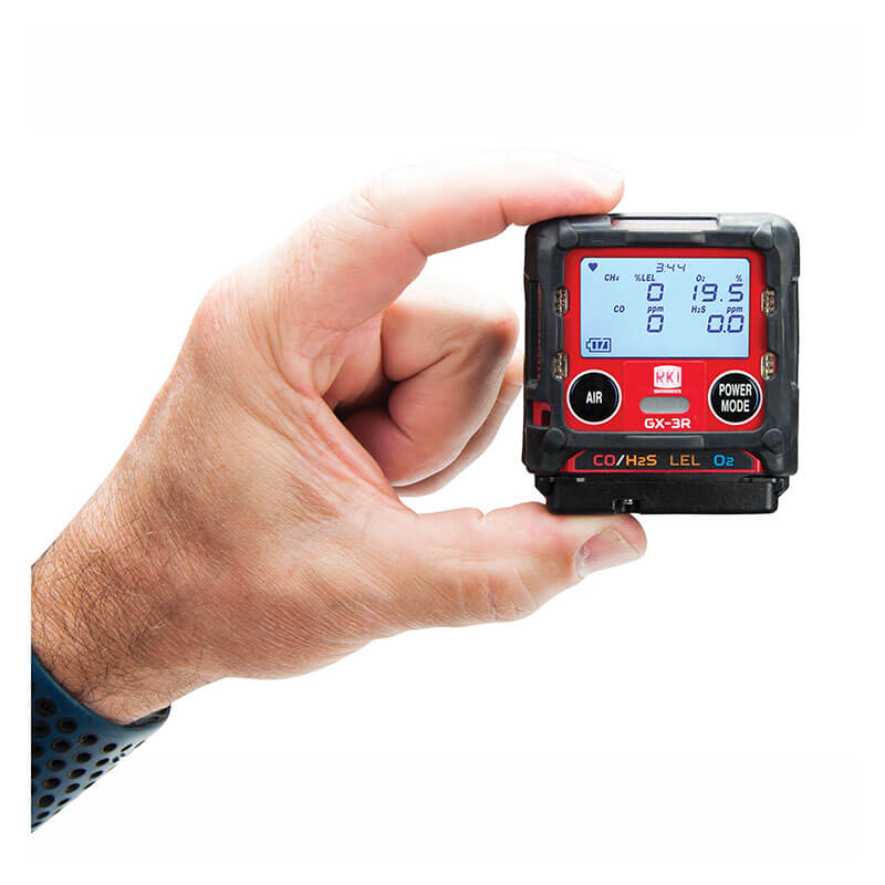 A hand holds the RKI Instruments GX-3R 4 Gas Confined Space Monitor 72-RA-C, a compact black device with "Air" and "Power Mode" buttons and a digital display for gas readings, ideal for confined space safety checks.
