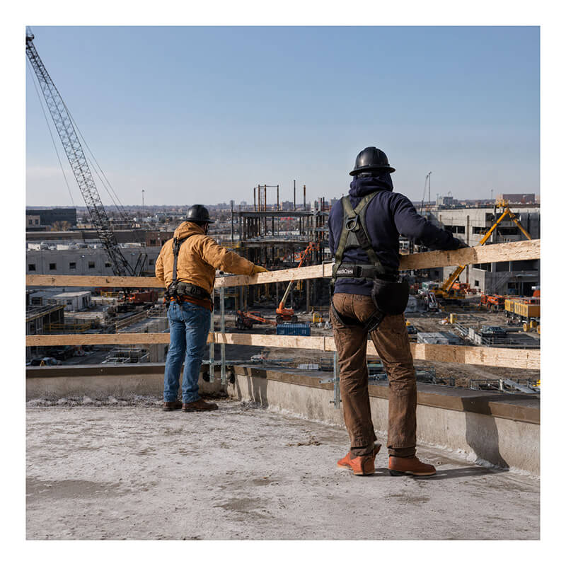 Two construction workers equipped with safety harnesses and hard hats stand on a rooftop, surveying an active construction site filled with cranes, scaffolding, and building materials beneath a clear sky. The French Creek Production Portable Guardrail System GR200 is installed along the roof edge, underscoring their dedication to fall prevention on the jobsite.