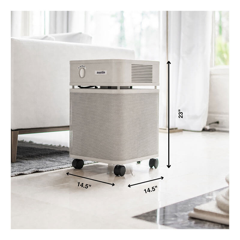 A white Austin Air Systems HealthMate Plus Machine B450 air purifier with wheels sits on a shiny floor by a sofa. It measures 23" tall and 14.5" wide/deep, featuring medical-grade HEPA filtration for formaldehyde removal.